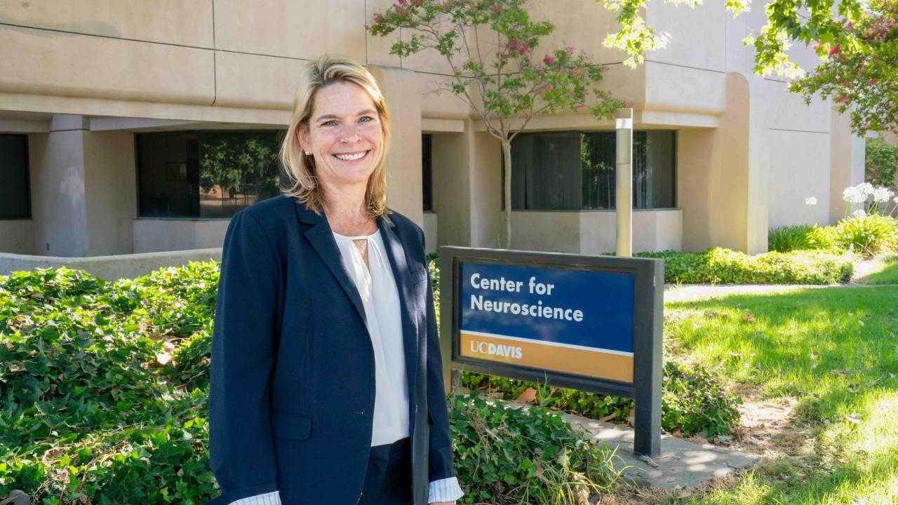 A woman in a navy suit stands smiling outside the UC Davis Center for Neuroscience building, next to a sign that reads “Center for Neuroscience UC Davis.” Green shrubs and trees surround the area.