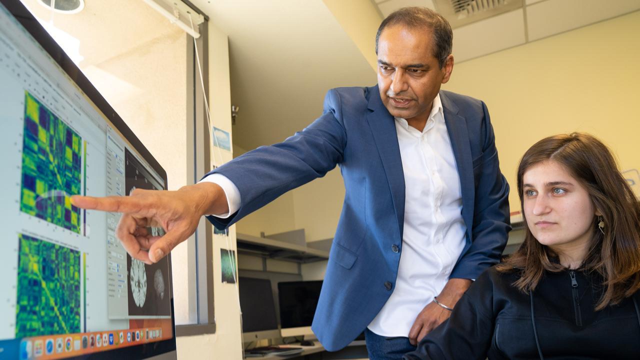 A male professor is standing and pointing at an image of a brain on a computer screen; a female trainee is seated in front of the computer.