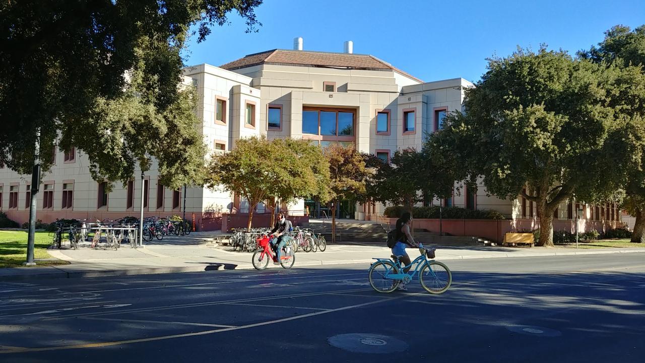 A modern beige-colored academic building with a red-tiled roof, large windows, and a symmetrical facade, located on a university campus. In the foreground, two people are riding bicycles—one on a red bike and the other on a blue bike. Several parked bicycles are visible near the building entrance, along with trees providing shade. The scene is illuminated by bright sunlight, casting shadows on the ground.