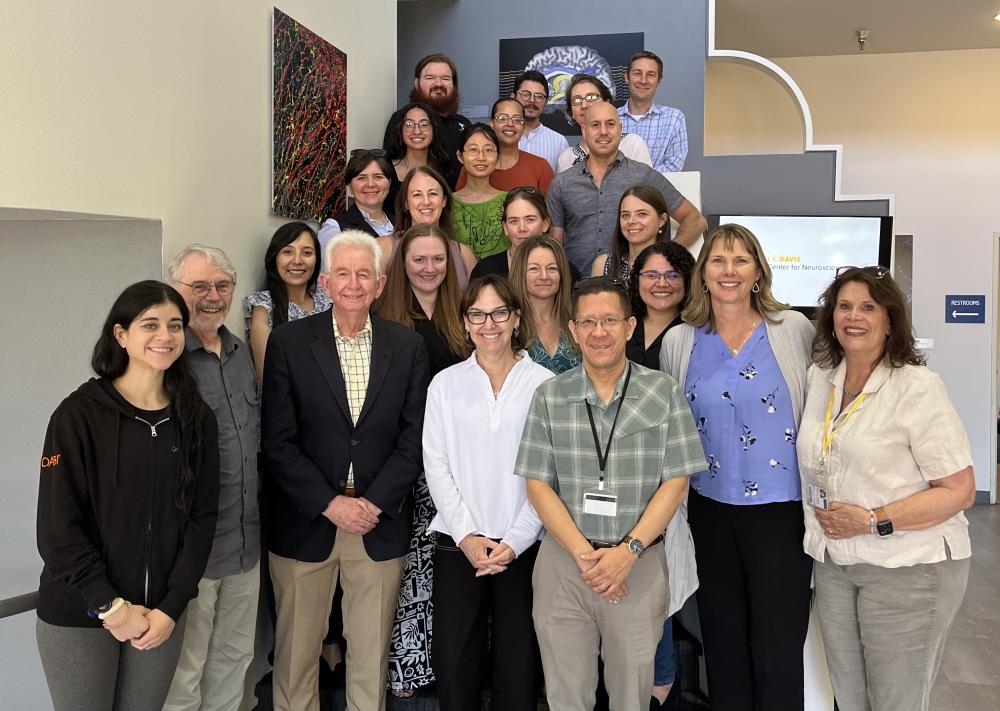 A large group of individuals posing for a group photo in an indoor setting. The group consists of men and women of varying ages, smiling and standing closely together. Behind them, neuroscience-themed artwork featuring neural imagery and a brain illustration is displayed on the wall. A screen in the background reads "UC Davis Center for Neuroscience," and a restroom sign is visible to the right. The setting is well-lit with light-colored walls and architectural detailing.